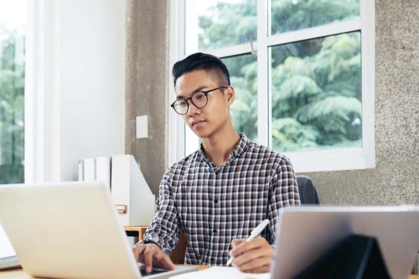 Student Working On Laptop Young serious Asian man in checked shirt and glasses reading information on laptop and making notes while sitting at table